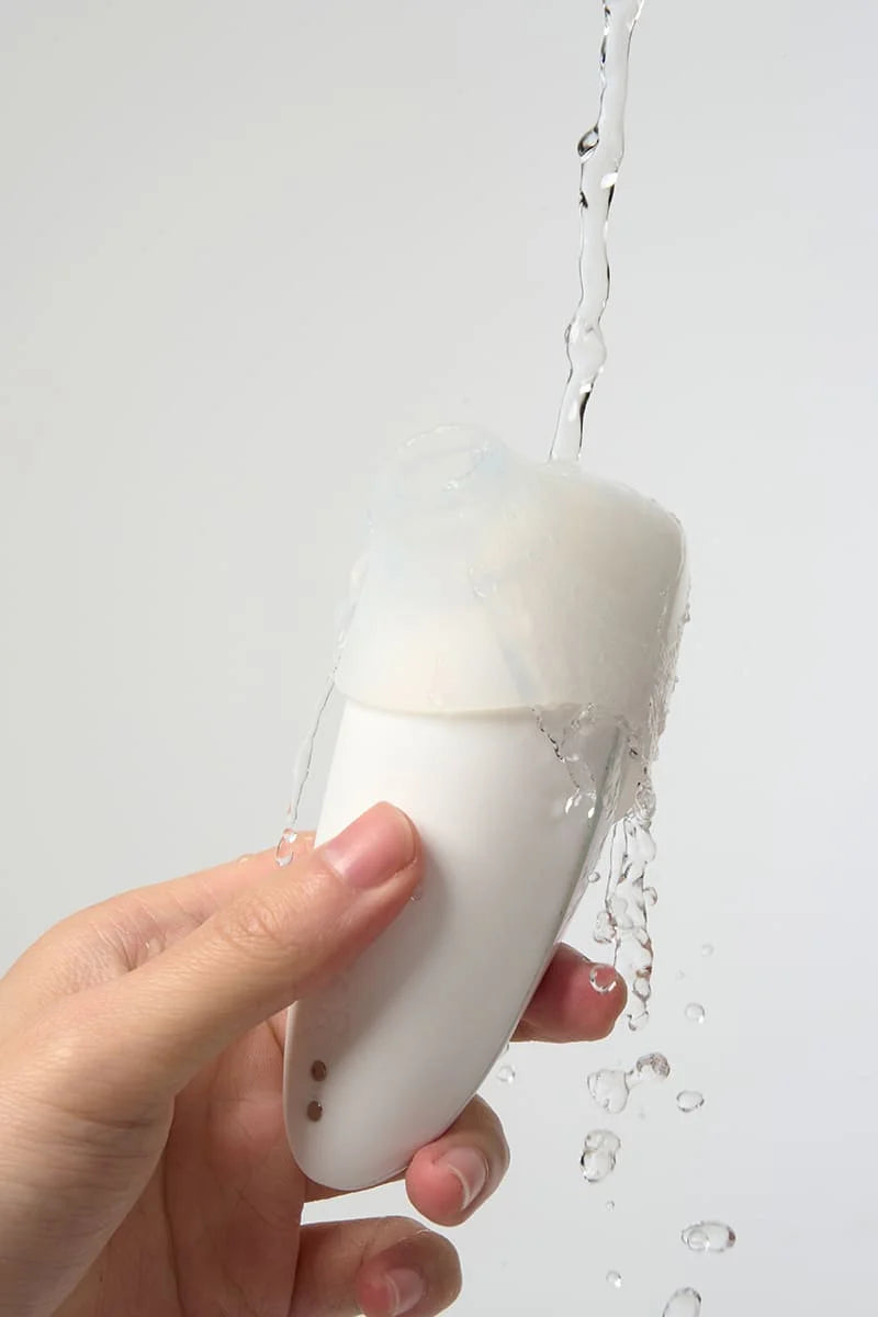 Hand holding a white Myvelvettouch Colorful Clitoral Slapping Massager with water being poured over it against a light gray background