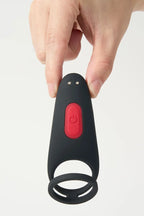 Hand holding a black and red Myvelvettouch Couples Vibrating Ring with Remote Control with a circular component on a light gray background